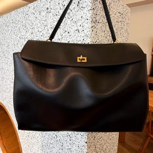 May include: A large, black handbag with a rectangular shape and a flap closure. The bag features a gold-colored clasp and a long, black handle. The bag is made of a smooth material and appears to be empty.