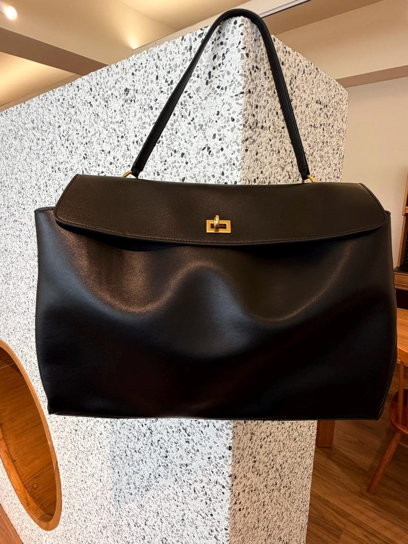 May include: A large, black handbag with a rectangular shape and a flap closure. The bag features a gold-colored clasp and a long, black handle. The bag is made of a smooth material and appears to be empty.