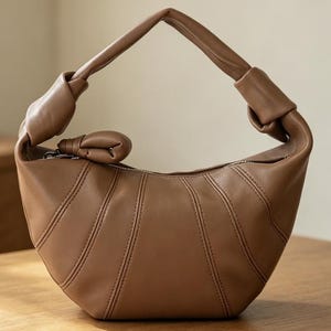 May include: A crescent-shaped handbag in a solid tan color. The bag features a smooth, matte finish with vertical stitched paneling, a single integrated handle with knotted ends, and a small knotted accent attached to the top zipper closure.