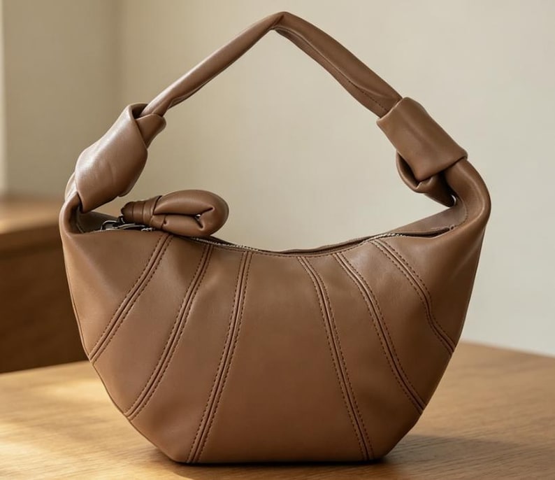 May include: A crescent-shaped handbag in a solid tan color. The bag features a smooth, matte finish with vertical stitched paneling, a single integrated handle with knotted ends, and a small knotted accent attached to the top zipper closure.