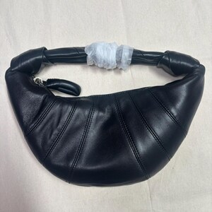 May include: A black crescent-shaped shoulder bag crafted from smooth leather. The bag features a curved silhouette with vertical stitched paneling, a knotted handle detail, and a top zipper closure with a leather pull. A white protective wrap covers the center of the handle.