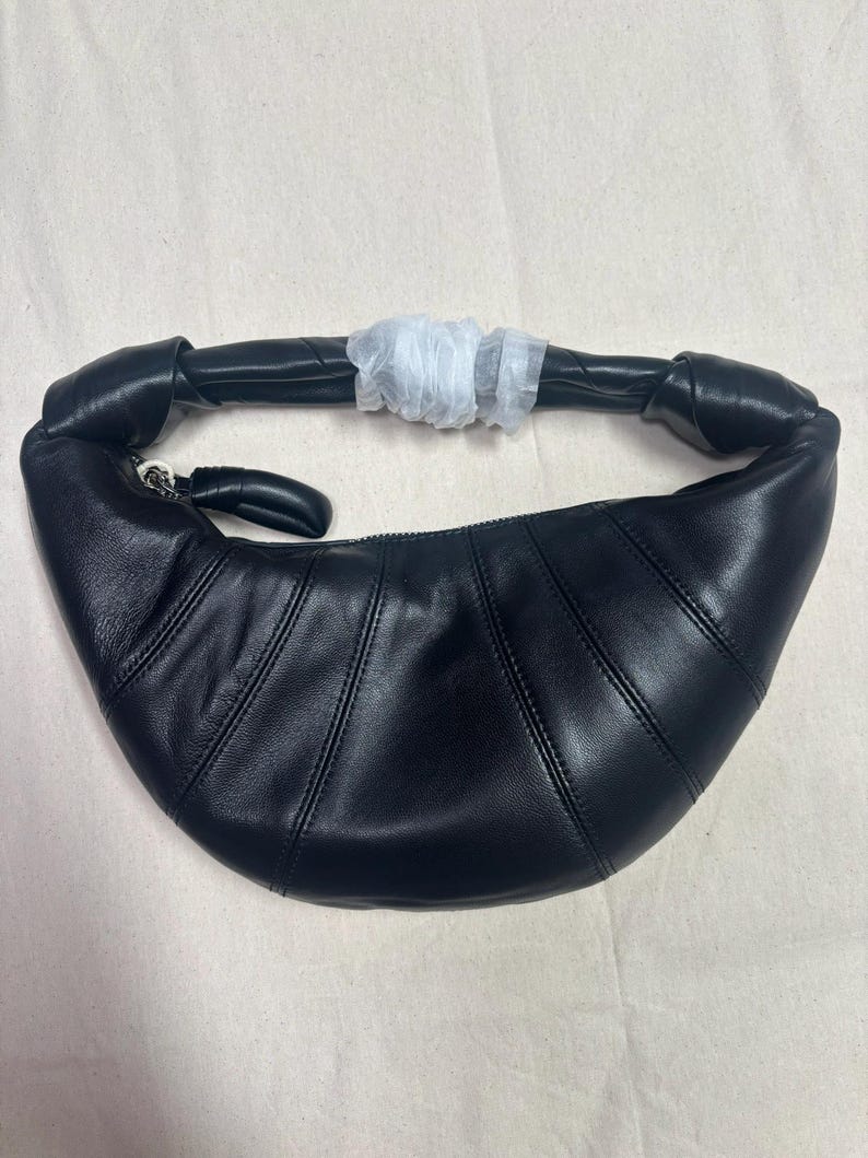 May include: A black crescent-shaped shoulder bag crafted from smooth leather. The bag features a curved silhouette with vertical stitched paneling, a knotted handle detail, and a top zipper closure with a leather pull. A white protective wrap covers the center of the handle.