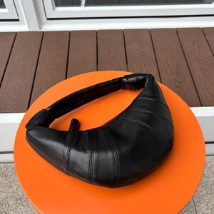 May include: A black crescent-shaped shoulder bag with a smooth, paneled leather-like texture. The bag features a curved silhouette, a thick integrated handle with knotted ends, and visible stitching along the panels, creating a structured look.