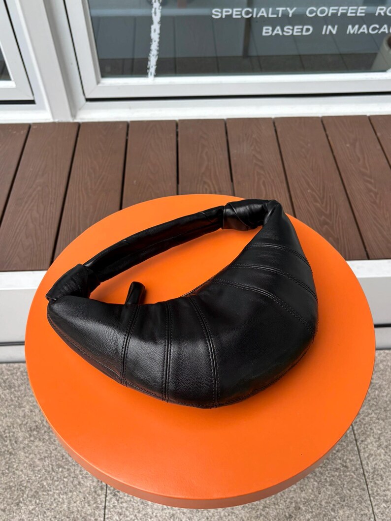 May include: A black crescent-shaped shoulder bag with a smooth, paneled leather-like texture. The bag features a curved silhouette, a thick integrated handle with knotted ends, and visible stitching along the panels, creating a structured look.