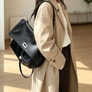 May include: A black leather shoulder bag with a pebbled texture, a fold-over flap, and a rectangular silver-tone metal clasp. The bag features a thin black shoulder strap and is worn over a beige trench coat, a white top, and dark trousers.