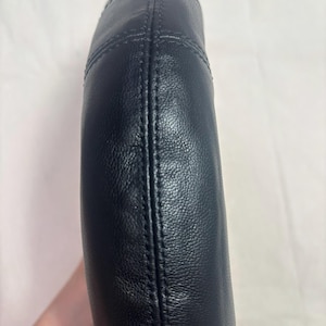 May include: A curved, black leather handle featuring a textured surface and prominent vertical stitching along the center. The handle has a rounded, padded construction with visible seams that run along the length of the piece, creating a structured look.