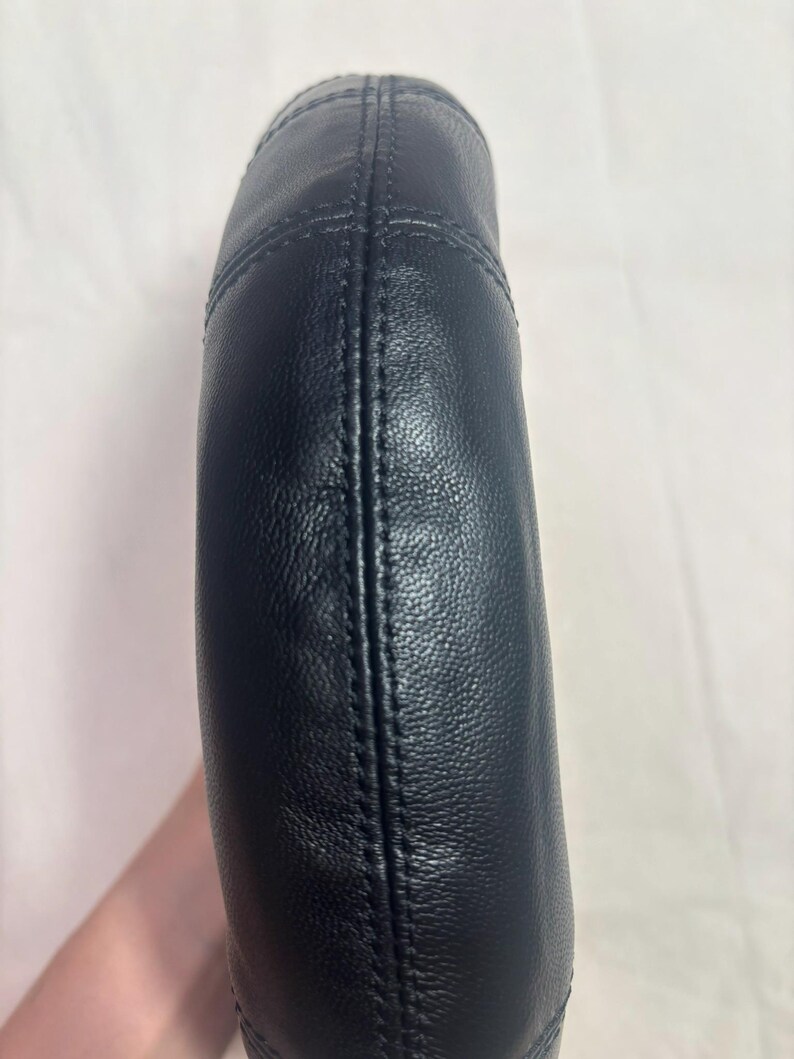 May include: A curved, black leather handle featuring a textured surface and prominent vertical stitching along the center. The handle has a rounded, padded construction with visible seams that run along the length of the piece, creating a structured look.