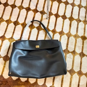 May include: A black leather handbag with a flap closure and a gold-colored clasp. The bag has a top handle and is set against a patterned brown and cream-colored surface.