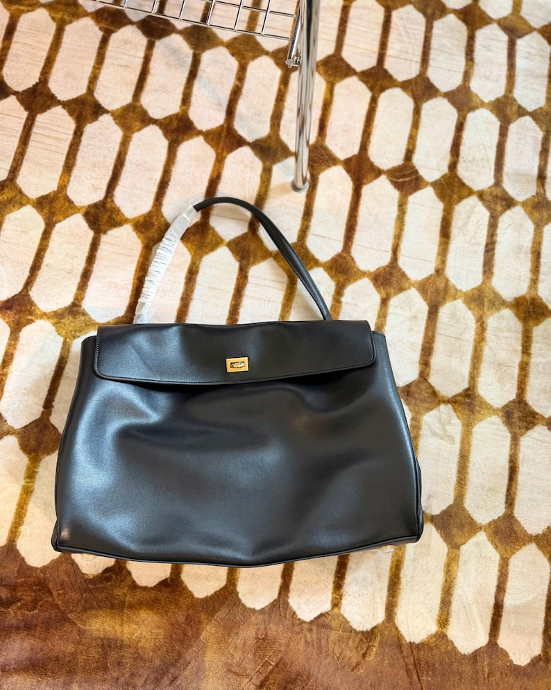 May include: A black leather handbag with a flap closure and a gold-colored clasp. The bag has a top handle and is set against a patterned brown and cream-colored surface.