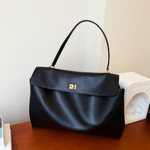 May include: A black leather handbag with a rectangular shape and a flap closure. The bag has a single handle and gold-colored hardware. The bag is sitting on a wooden surface.