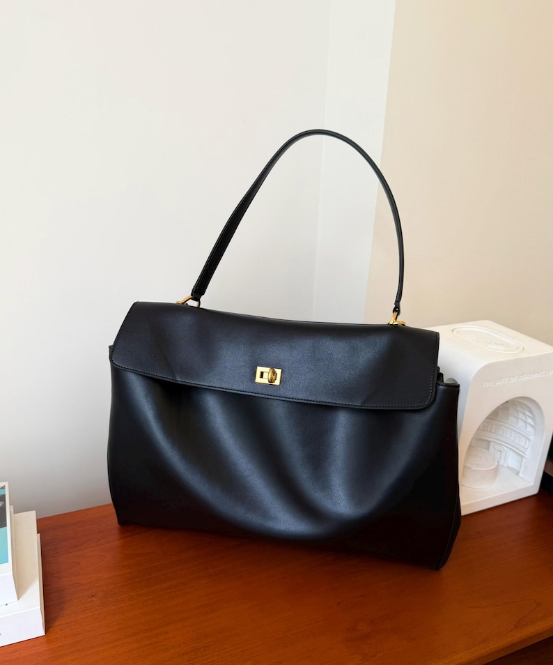 May include: A black leather handbag with a rectangular shape and a flap closure. The bag has a single handle and gold-colored hardware. The bag is sitting on a wooden surface.