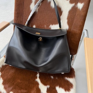 May include: A black leather handbag with a top handle and a gold-colored clasp. The bag has a rectangular shape with a flap closure. The bag is sitting on a chair with a brown and white cowhide pattern.