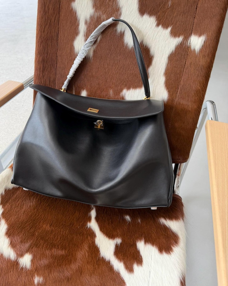 May include: A black leather handbag with a top handle and a gold-colored clasp. The bag has a rectangular shape with a flap closure. The bag is sitting on a chair with a brown and white cowhide pattern.