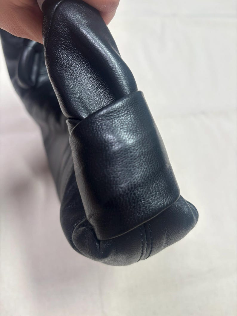 May include: A black leather handbag featuring a soft, gathered, and pleated texture. The bag has a smooth, rounded side panel with visible stitching along the bottom edge, showcasing a flexible and slouchy construction with a matte finish.