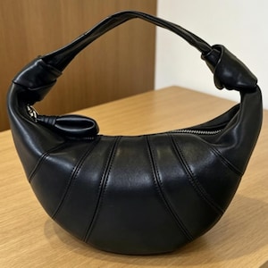 May include: A black crescent-shaped handbag with a smooth finish and vertical stitched paneling. The bag features a single integrated handle with a knotted detail on each side and a silver-toned zipper closure along the top curved edge.