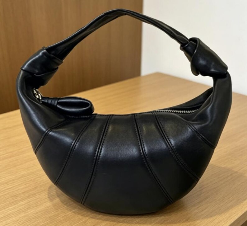 May include: A black crescent-shaped handbag with a smooth finish and vertical stitched paneling. The bag features a single integrated handle with a knotted detail on each side and a silver-toned zipper closure along the top curved edge.