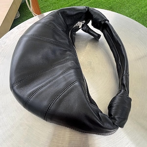 May include: A black leather crescent-shaped shoulder bag featuring a curved silhouette with vertical stitched panels, a silver-toned metal zipper closure along the top edge, and a knotted handle detail on one side of the bag's structure.