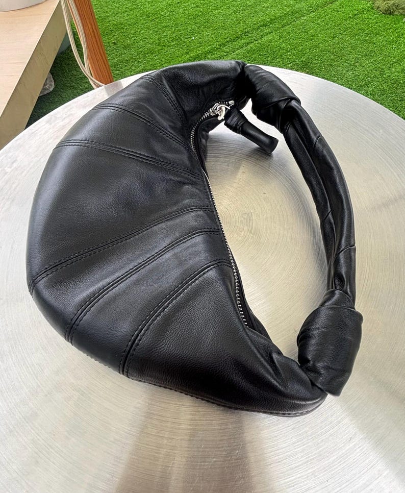May include: A black leather crescent-shaped shoulder bag featuring a curved silhouette with vertical stitched panels, a silver-toned metal zipper closure along the top edge, and a knotted handle detail on one side of the bag's structure.
