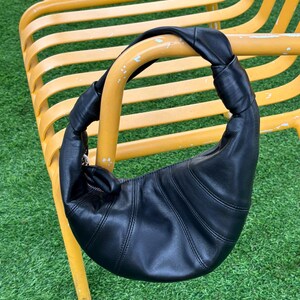 May include: A black leather crescent-shaped shoulder bag featuring a knotted handle design and a curved silhouette with visible stitched paneling. The bag includes a silver-toned metal zipper closure along the top edge, providing a secure opening.