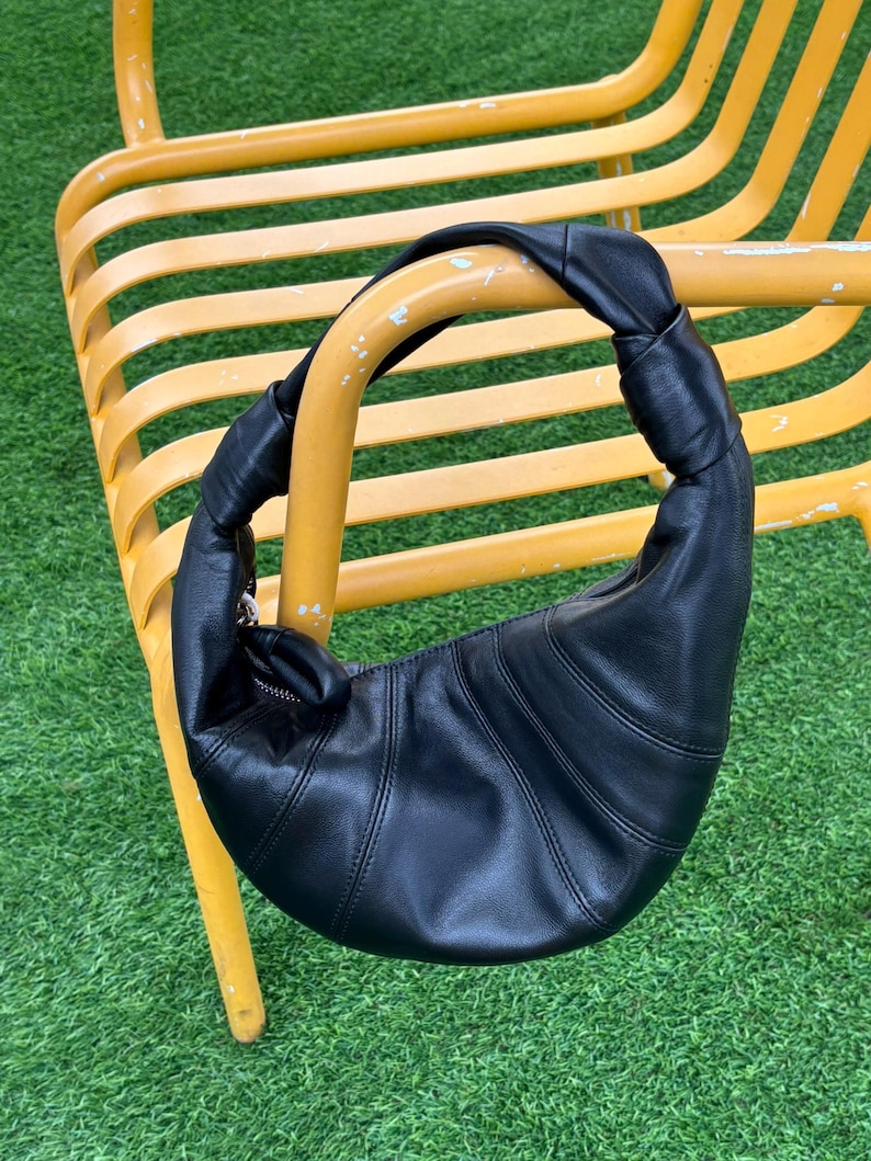 May include: A black leather crescent-shaped shoulder bag featuring a knotted handle design and a curved silhouette with visible stitched paneling. The bag includes a silver-toned metal zipper closure along the top edge, providing a secure opening.