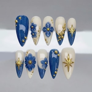 May include: A set of ten artificial nails with a variety of designs. Some nails feature a denim-like blue base, while others have a white base. Embellishments include gold starbursts, gold beads, and blue floral accents. The nails are almond-shaped.