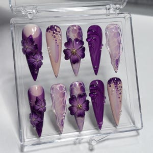 May include: A set of ten artificial nails in a clear plastic case. The nails feature a variety of designs, including floral embellishments, glitter, and abstract patterns. The color palette includes shades of purple, lavender, and nude.