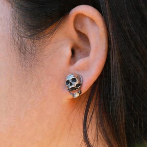 May include: A silver-toned stud earring shaped like a detailed human skull with dark, recessed eye sockets and a defined jawline. The metallic surface features a polished finish with darkened accents in the crevices to highlight the anatomical features.