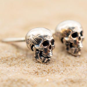 May include: Two silver skull-shaped earrings resting on sand. The earrings have detailed facial features, including eye sockets, a nose cavity, and a jawline. The skulls are slightly weathered, giving them a vintage appearance. The background is a sandy beach.