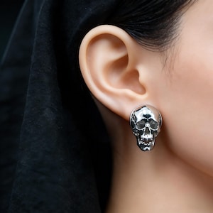 Può includere: Un orecchino a lobo in metallo color argento lucido a forma di teschio umano. L'orecchino presenta una finitura metallica altamente riflettente con dettagliati contorni anatomici delle orbite, della cavità nasale e dei denti.