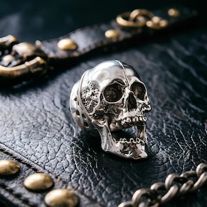 Può includere: Un anello in argento lucido a forma di teschio umano dettagliato con la mascella aperta, caratterizzato da complesse texture ossee e superfici lisce, esposto accanto a un cinturino in pelle nera con borchie in metallo color ottone e fibbia.