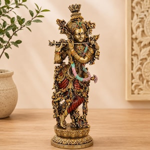 May include: A detailed bronze-toned statue of a deity playing a flute, standing on a lotus-shaped base. The figure wears ornate jewellery, a crown, and a red garment, with a multi-coloured floral garland draped around the neck and shoulders.