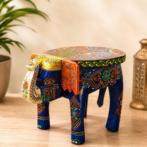 May include: A small, hand-painted wooden stool shaped like an elephant. The stool features a deep blue base color with intricate, multi-colored floral and geometric patterns in red, green, yellow, and white across the legs, ears, and circular top.