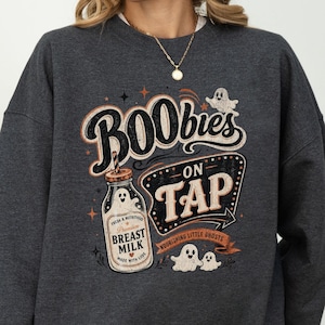 May include: A dark charcoal gray crewneck sweatshirt featuring a graphic print with the text BOObies ON TAP. The design includes a glass bottle labeled BREAST MILK, small ghost illustrations, and decorative stars in shades of cream and orange.