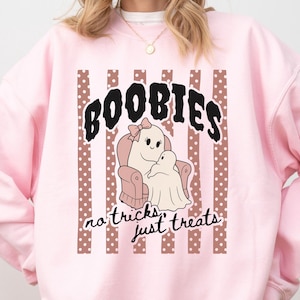 May include: A light pink crewneck sweatshirt featuring a graphic print of two ghosts sitting in a chair. The text reads BOOBIES and no tricks just treats. The design includes brown vertical stripes with white polka dots behind the ghosts.