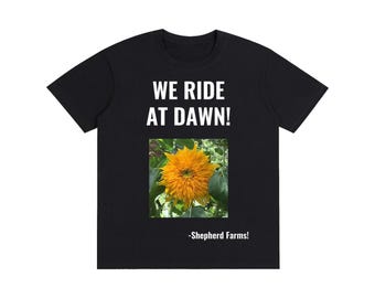 Può includere: Una t-shirt nera a maniche corte con il testo bianco WE RIDE AT DAWN! sopra una stampa fotografica quadrata di un vivace girasole giallo con foglie verdi. Il testo -Shepherd Farms! è stampato in bianco sotto l'immagine del girasole.