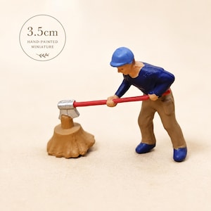 May include: Hand-painted miniature figurine of a lumberjack chopping wood. The figure is wearing a blue shirt, brown pants, and a blue hat. The axe has a red handle. The miniature is 3.5cm tall.