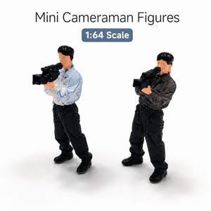 May include: Two 1:64 scale mini cameraman figures with the text Mini Cameraman Figures 1:64 Scale. One figure wears a light blue shirt and black pants, the other a dark gray shirt and black pants. Both hold black video cameras to their faces.