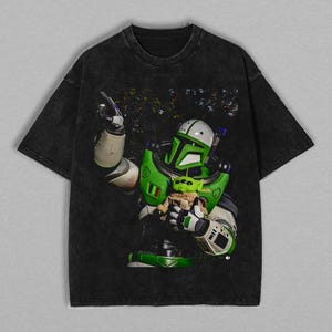 K&ouml;nnte beinhalten: Schwarzes T-Shirt mit einer Grafik eines gr&uuml;n-wei&szlig;en Weltraum-Rangers, der einen kleinen gr&uuml;nen Au&szlig;erirdischen h&auml;lt. Der Weltraum-Ranger tr&auml;gt einen gr&uuml;n-wei&szlig;en Anzug mit Helm. Der Au&szlig;erirdische ist in eine braune Decke geh&uuml;llt. Das T-Shirt hat einen verblichenen, Vintage-Look.