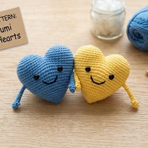 May include: A pair of crocheted hearts, one blue and one yellow, with smiling faces and attached limbs. The image includes yarn, a crochet hook, and a tag that reads "CROCHET PATTERN: Amigurumi Couple Hearts."
