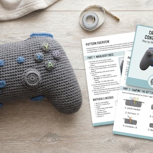 May include: A gray crocheted pillow shaped like a video game controller with blue accents and buttons labeled Y, X, B, and A. Beside it are printed instruction sheets titled Crochet Gamer Controller Pillow, along with yarn, a hook, and stitch markers.