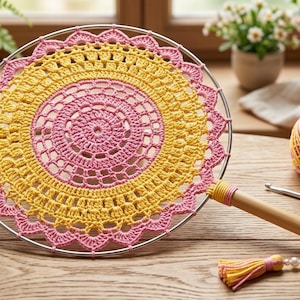 Könnte beinhalten: Ein runder Handfächer mit einer Häkeldecke in abwechselnden rosa und gelben Garnmustern, die über einen silbernen Metallring gespannt ist. Der Fächer hat einen Holzgriff, der mit passendem Garn umwickelt und mit einer Quaste und weißen Perlen verziert ist.