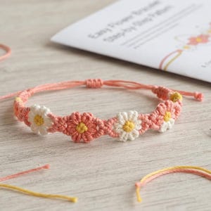 May include: A handmade macrame bracelet featuring a repeating pattern of small white and coral-colored flowers with yellow centers. The bracelet has adjustable sliding knots and is displayed alongside a printed instruction sheet, scissors, and loose thread.