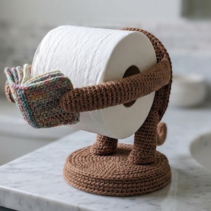 May include: A brown crocheted toilet roll holder shaped like a monkey. The holder has a circular base and arms that embrace the toilet roll. The monkey's hands are multi-coloured.