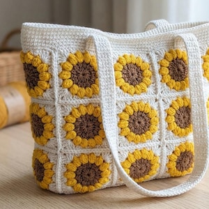 Puede incluir: Bolso tote de crochet blanco con diseño de girasoles amarillos y marrones. Tiene dos asas blancas y está sobre una superficie de madera.