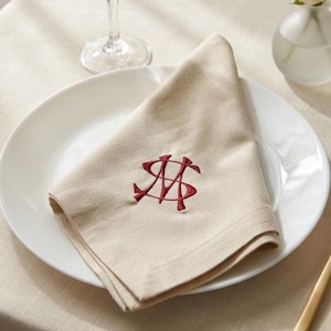Könnte beinhalten: Eine beigefarbene Stoffserviette, die zu einem Dreieck gefaltet auf einem weißen Teller liegt. Die Serviette ist mit einem Monogramm aus dunkelrotem Garn bestickt, das aus den ineinander verschlungenen Großbuchstaben S und M besteht.