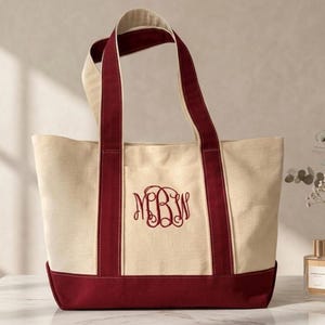 May include: A cream-colored canvas tote bag featuring deep burgundy accents on the base and the two vertical handles. The front of the bag displays an embroidered monogram with the letters MBW in a decorative, swirling script font in burgundy.