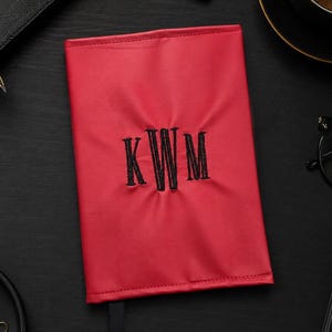May include: A red notebook cover with black embroidered initials "KWM" is centered on a black surface. A black ribbon bookmark extends from the cover. The scene includes a pen, glasses, a cup of coffee, and other office supplies.