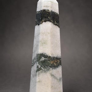 May include: A polished stone obelisk featuring a pointed top and a multi-faceted vertical body. The stone is primarily white with prominent, irregular horizontal bands and patches of dark forest green and charcoal gray mineral inclusions throughout.
