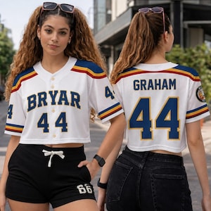 May include: White baseball-style crop top with blue and gold accents. The front displays "BRYAR 44" in blue lettering, while the back shows "GRAHAM 44". The sleeves and shoulders have blue, gold, and red stripes. The model is wearing black shorts.