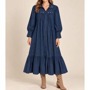 May include: A dark blue denim midi dress featuring a button-down front, long sleeves with cuffed wrists, and a gathered tiered skirt. The collar area is accented with small white studs, and the fabric has a visible denim texture throughout.
